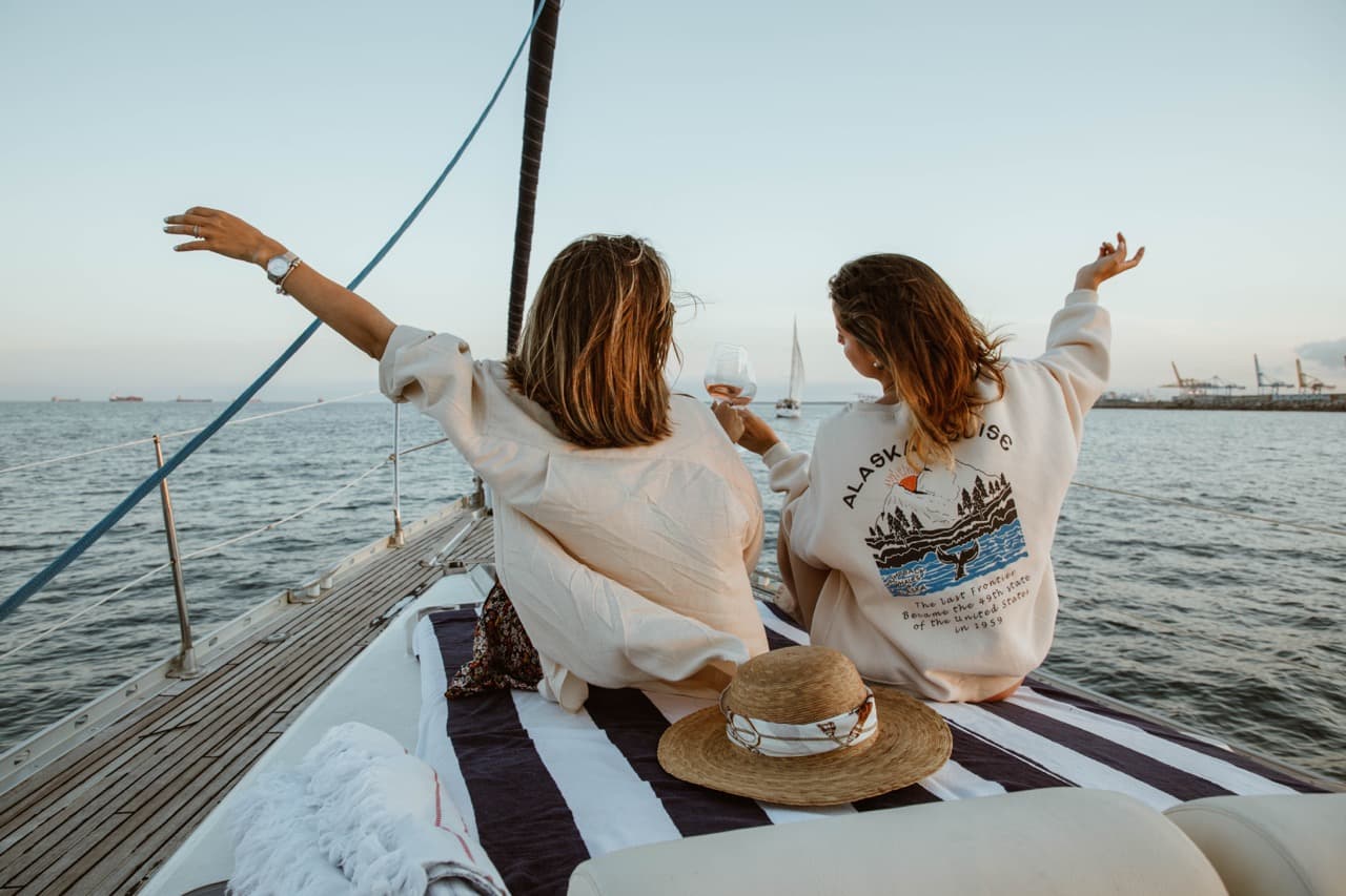 Dos amigas con los brazos alzados disfrutando de la proa de un velero al atardecer