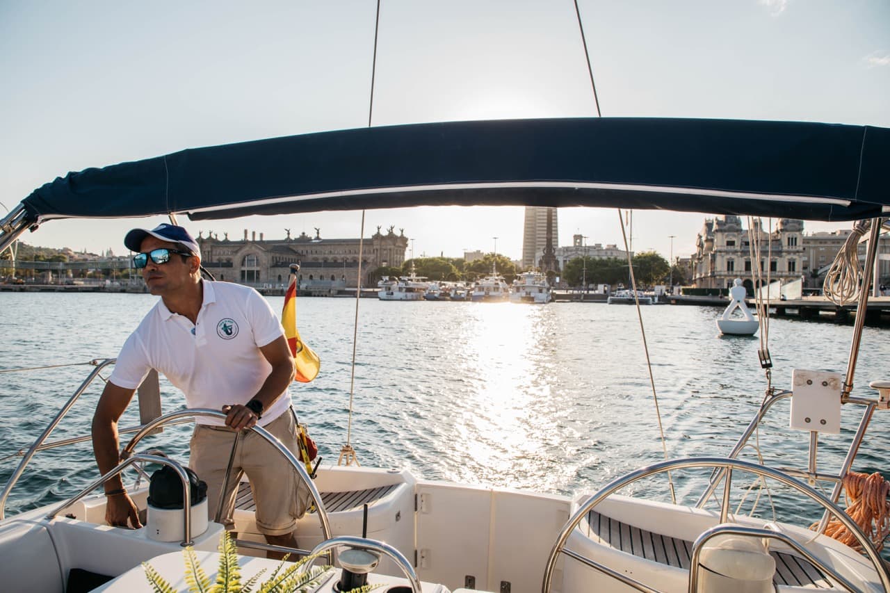 Patrón al timón de un velero con el Port Vell de Barcelona iluminado al atardecer