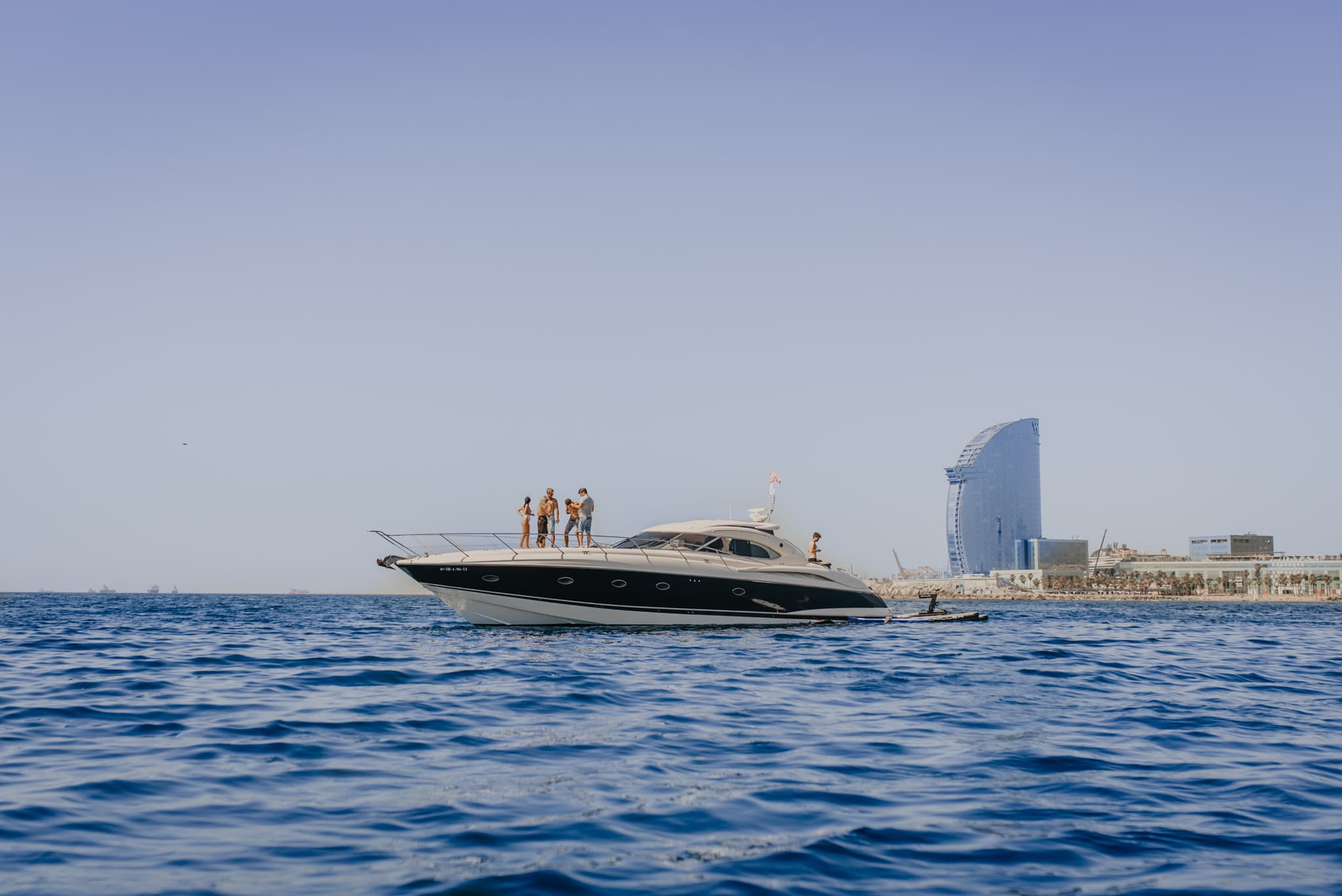 Yate fondeado frente a la costa de Barcelona con el Hotel W visible y pasajeros en la proa