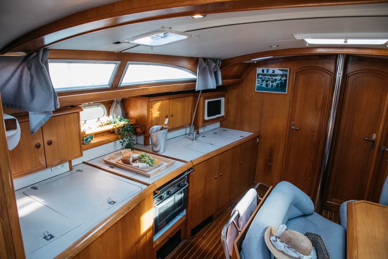 Interior de camarote de velero con cocina de madera, mesa con queso, uvas y pan, y sombrero sobre el asiento