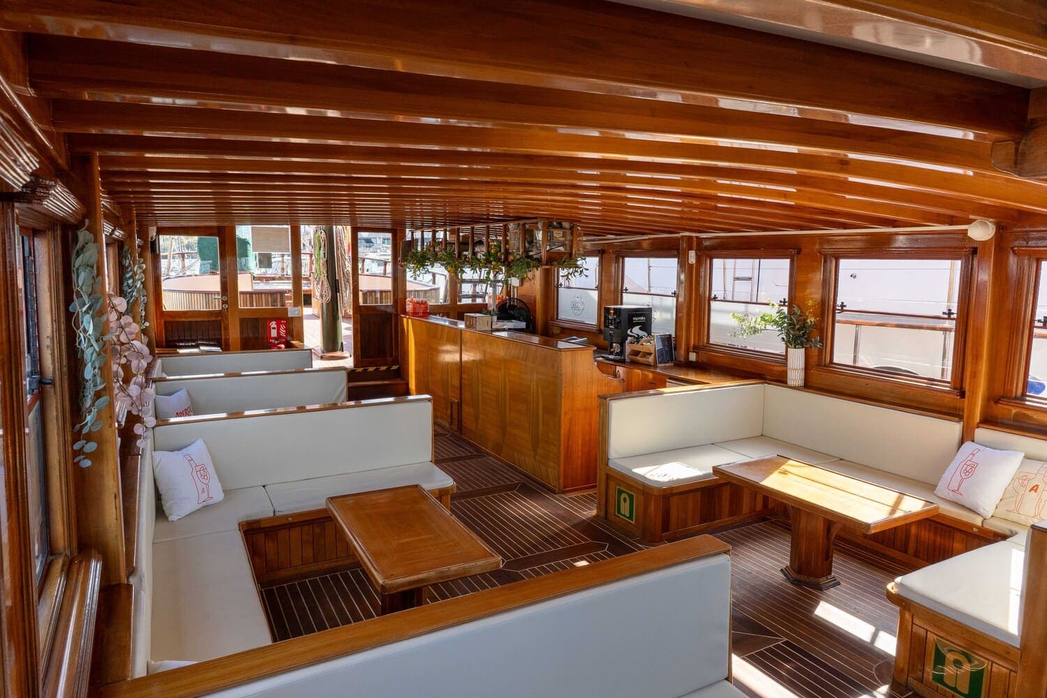 Salón interior de madera de un velero con bancos blancos y barra, con vistas al puerto