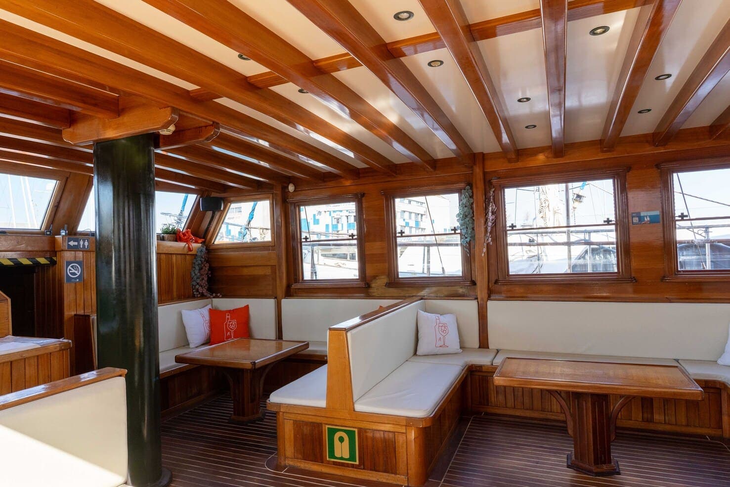 Salón interior de madera de un velero con bancos blancos y cojines junto a la columna del timón