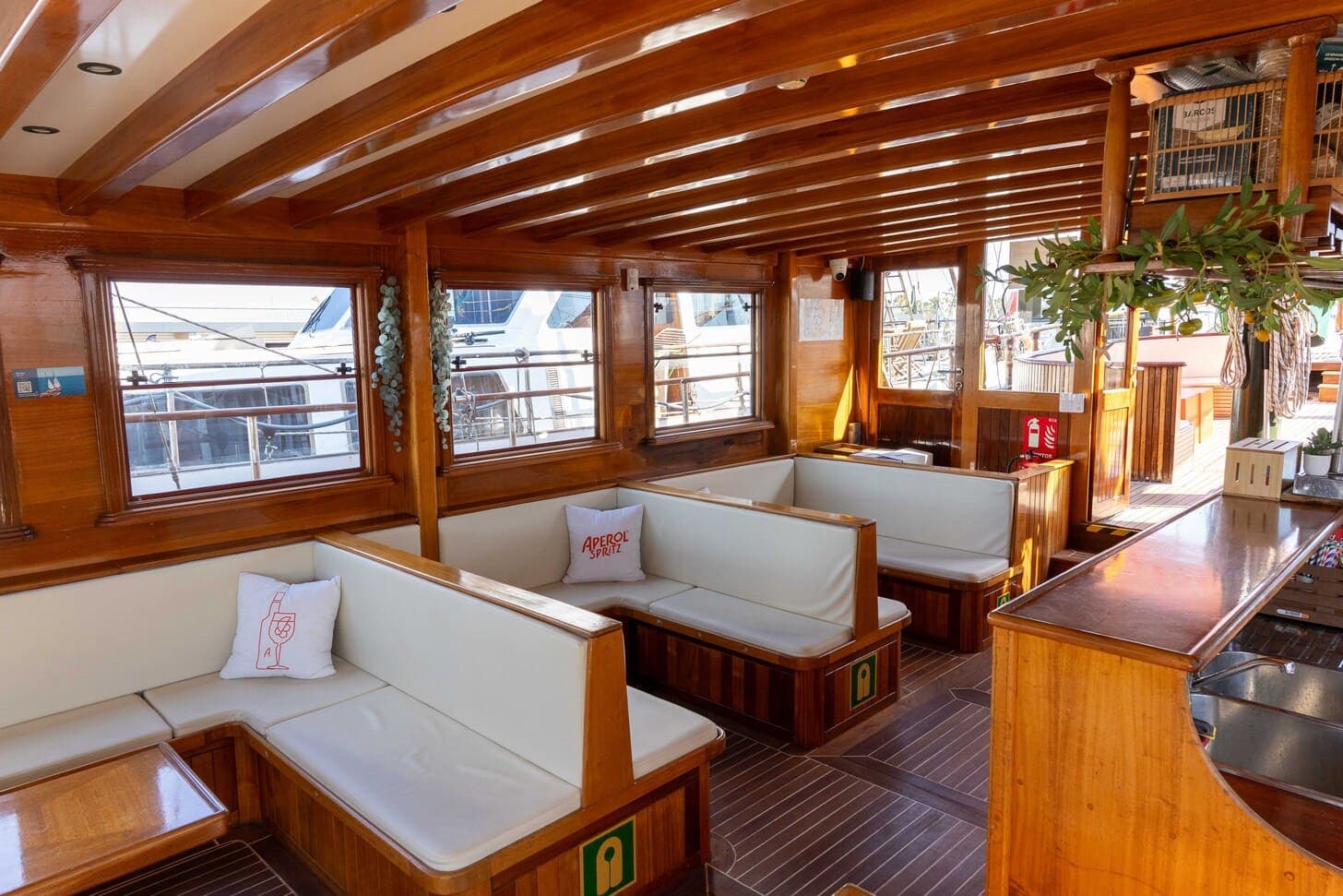Salón interior de madera de un velero con bancos blancos, barra y ramas de cítricos colgando