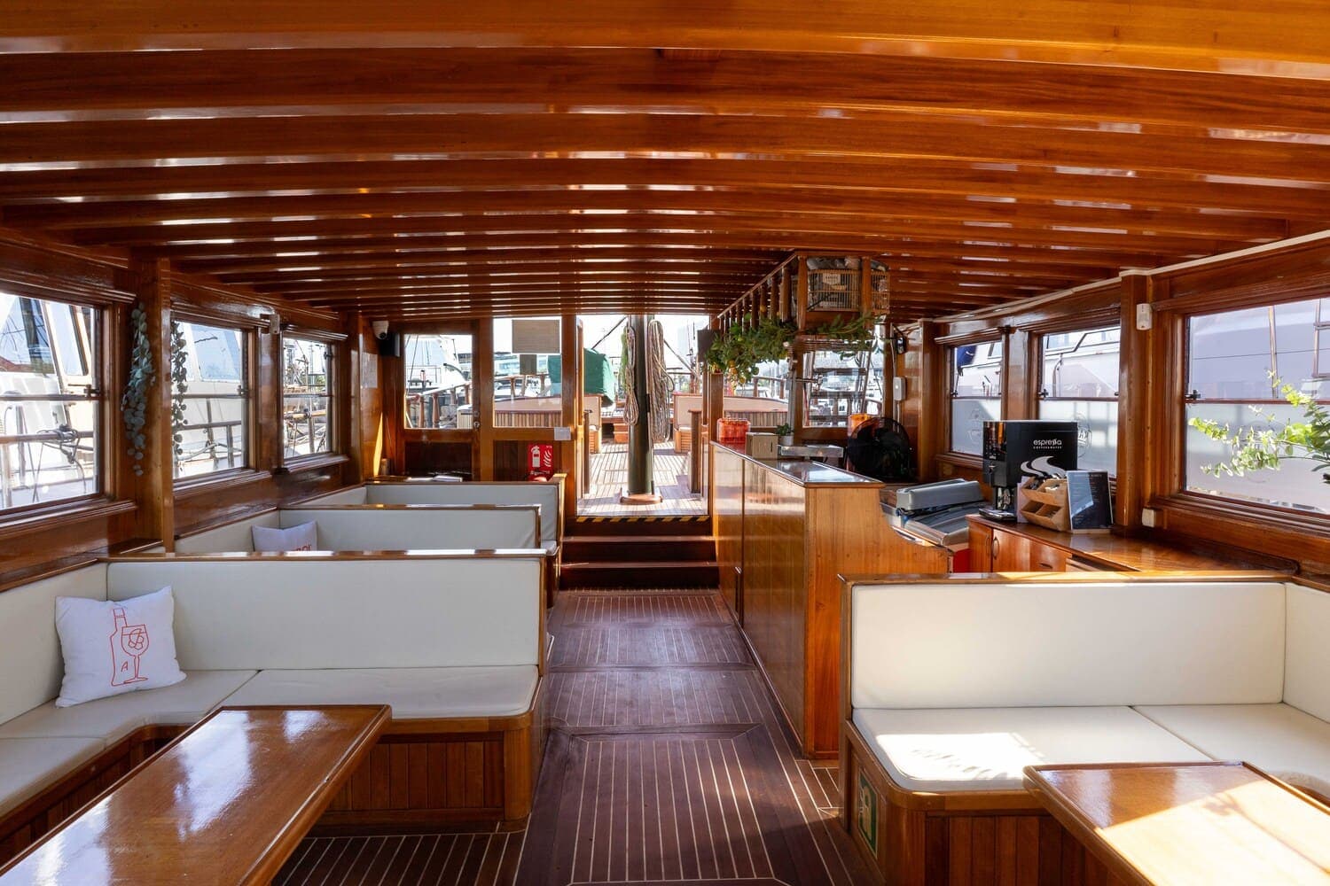 Salón interior de madera de un velero con bancos blancos y barra al fondo