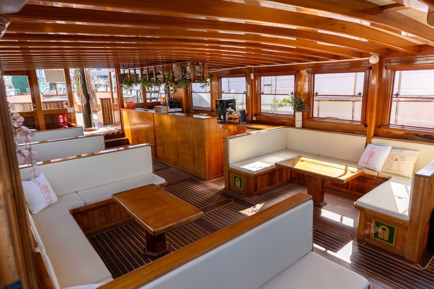 Salón interior de madera de un velero con bancos blancos, mesas y barra al fondo
