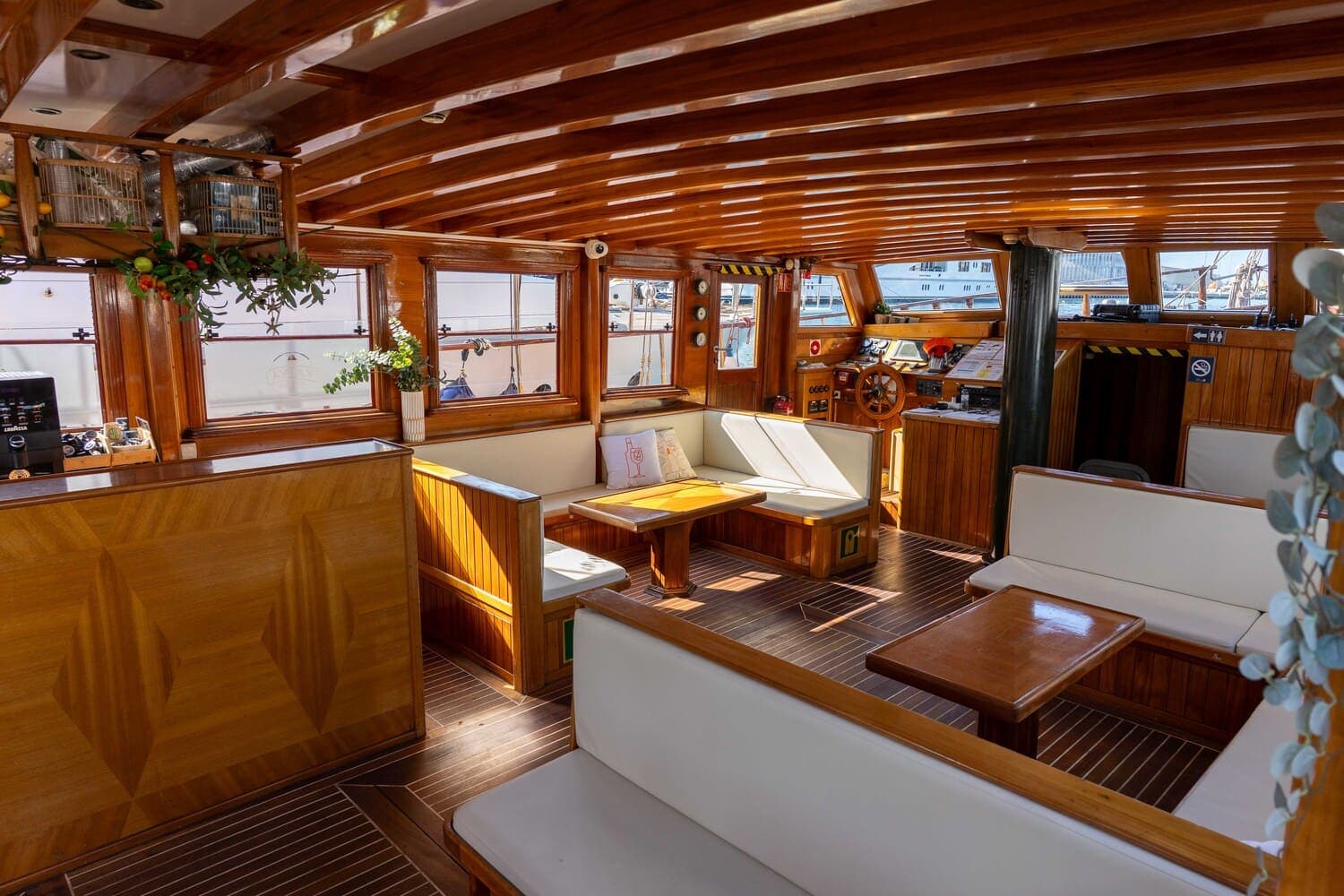 Salón interior de madera de un velero con bancos blancos junto al timón y el puesto de mando