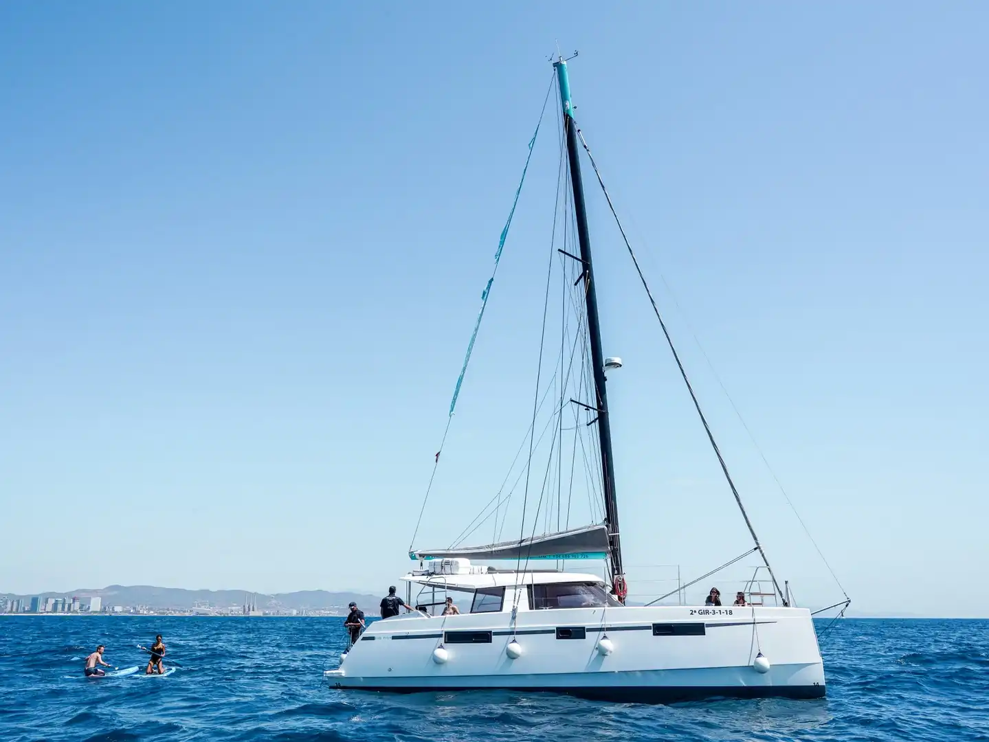 Catamarán de vela fondeado con gente a bordo y dos personas haciendo paddle surf junto al casco
