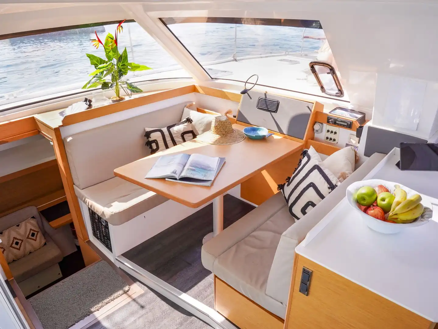 Salón comedor interior de catamarán con mesa de madera, bancos y frutero, con vistas al mar