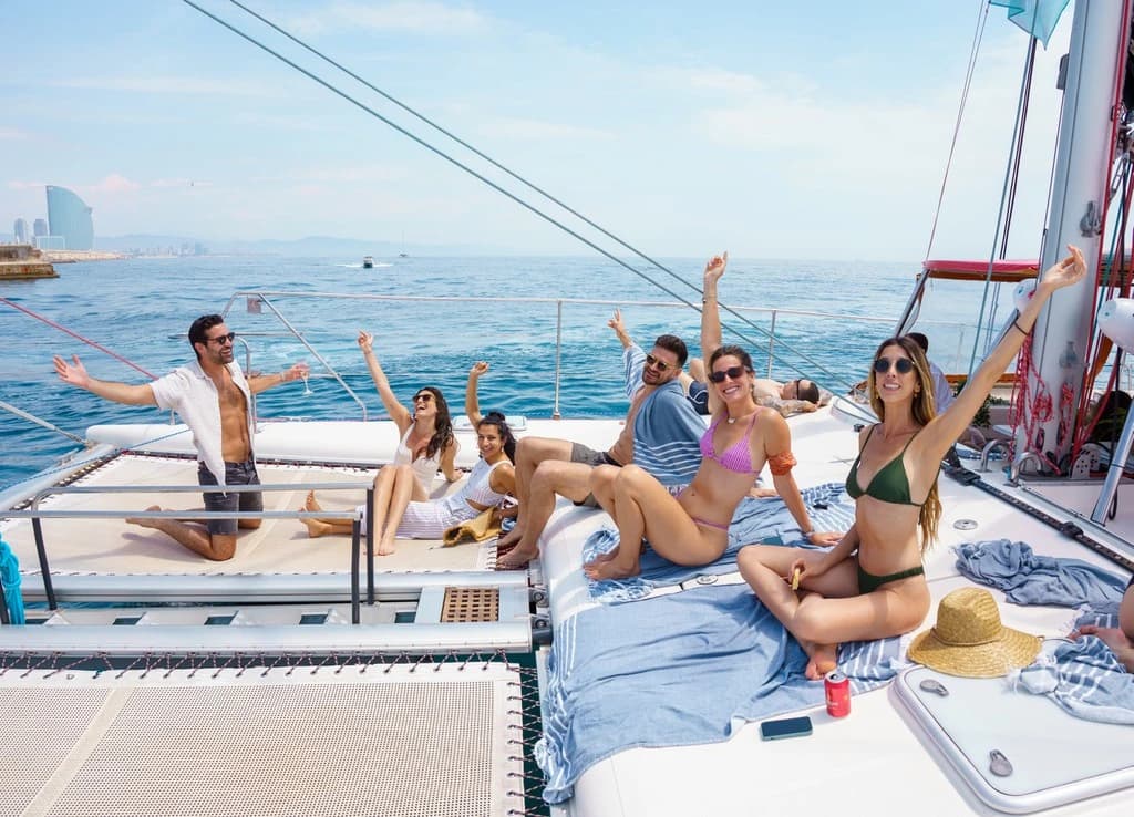 Amigos brindando con los brazos en alto sentados en la proa de un catamarán, con skyline al fondo