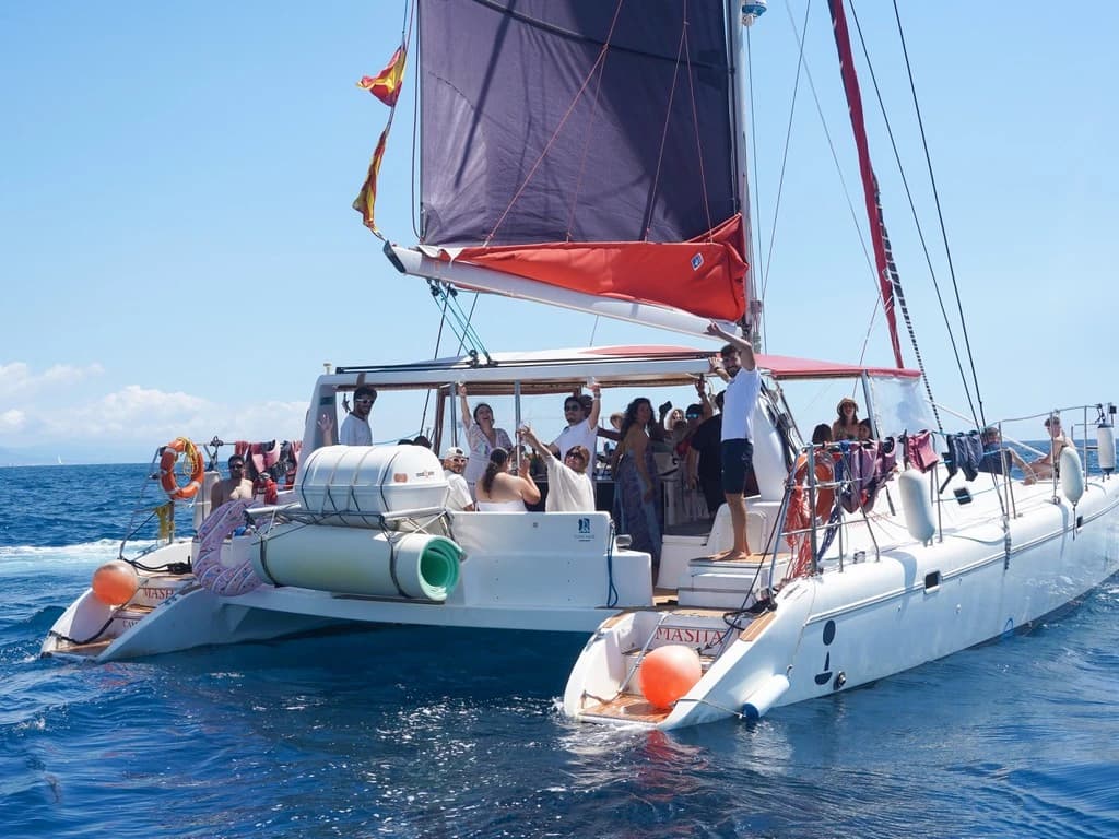 Gran catamarán de vela con pasajeros brindando a bordo, velas recogidas y defensas naranjas