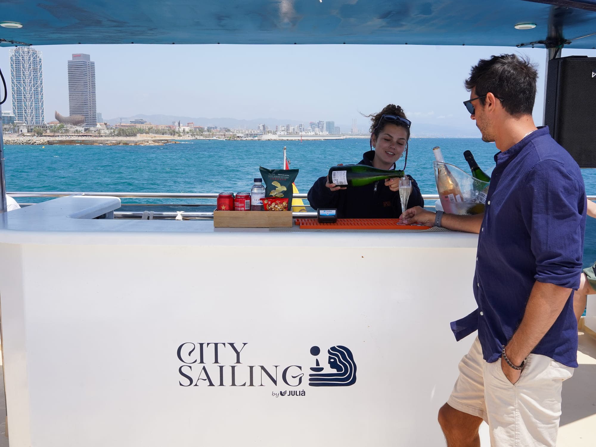 Tripulante sirviendo cava a un pasajero en la barra a bordo del catamarán City Sailing