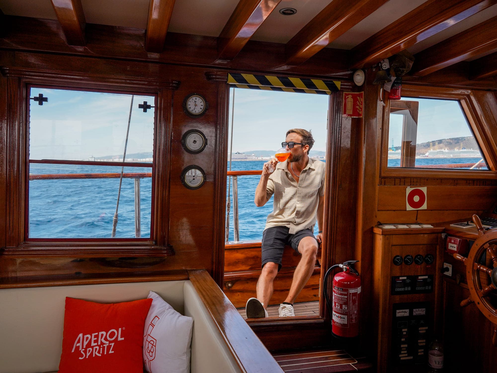 Hombre bebiendo un Aperol Spritz junto al timón de madera del barco con el mar de fondo