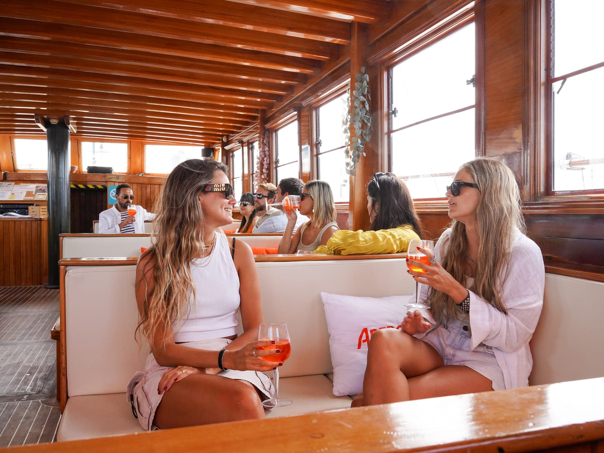 Amigas conversando con copas de Aperol Spritz en el salón interior de madera del barco