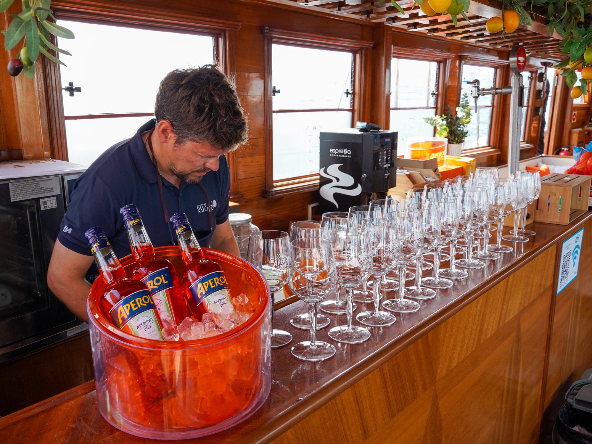 Tripulante preparando botellas de Aperol en hielo junto a una fila de copas en la barra del barco