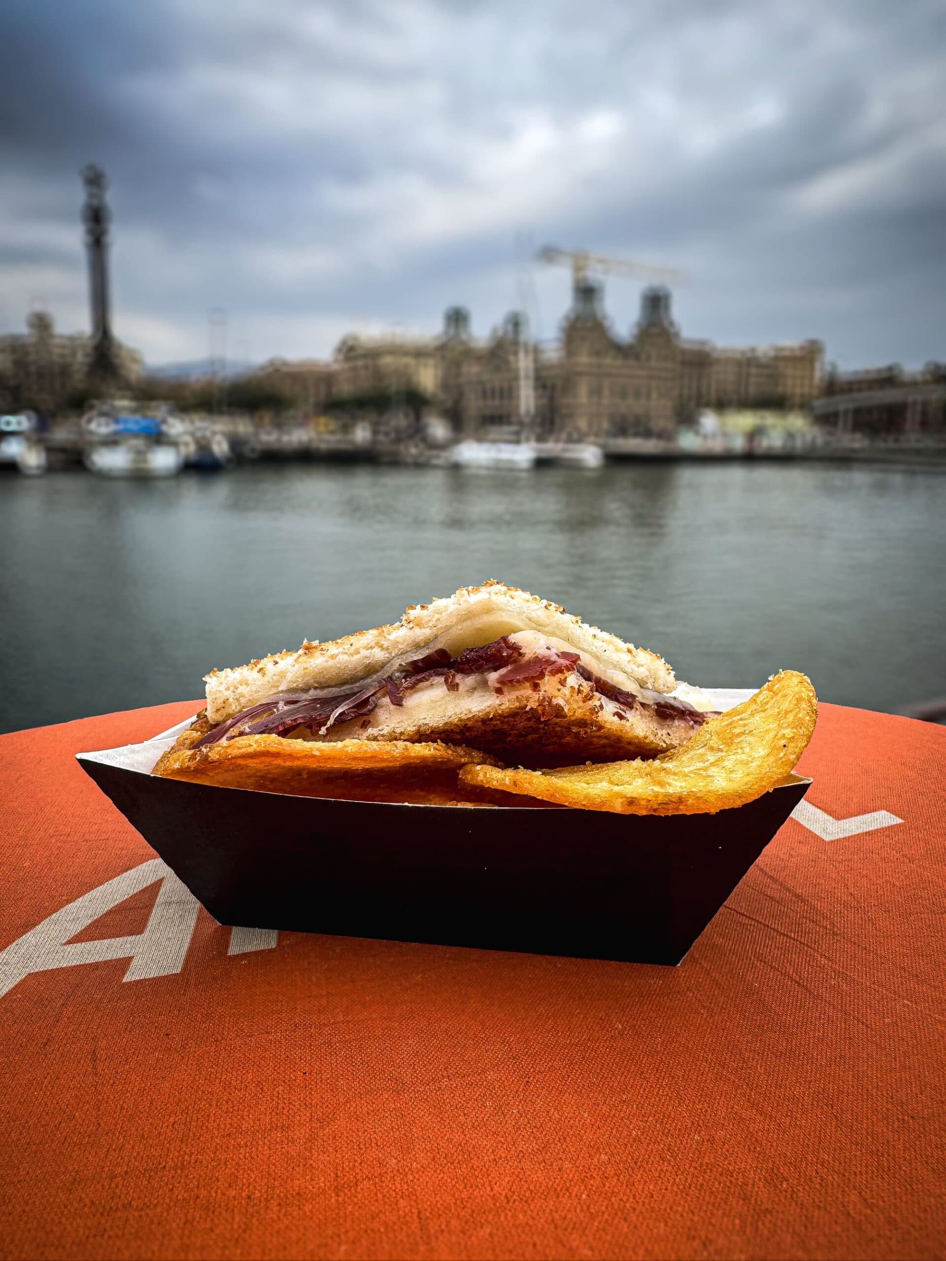 Bocadillo de jamón crujiente en bandeja de cartón con el puerto de Barcelona de fondo
