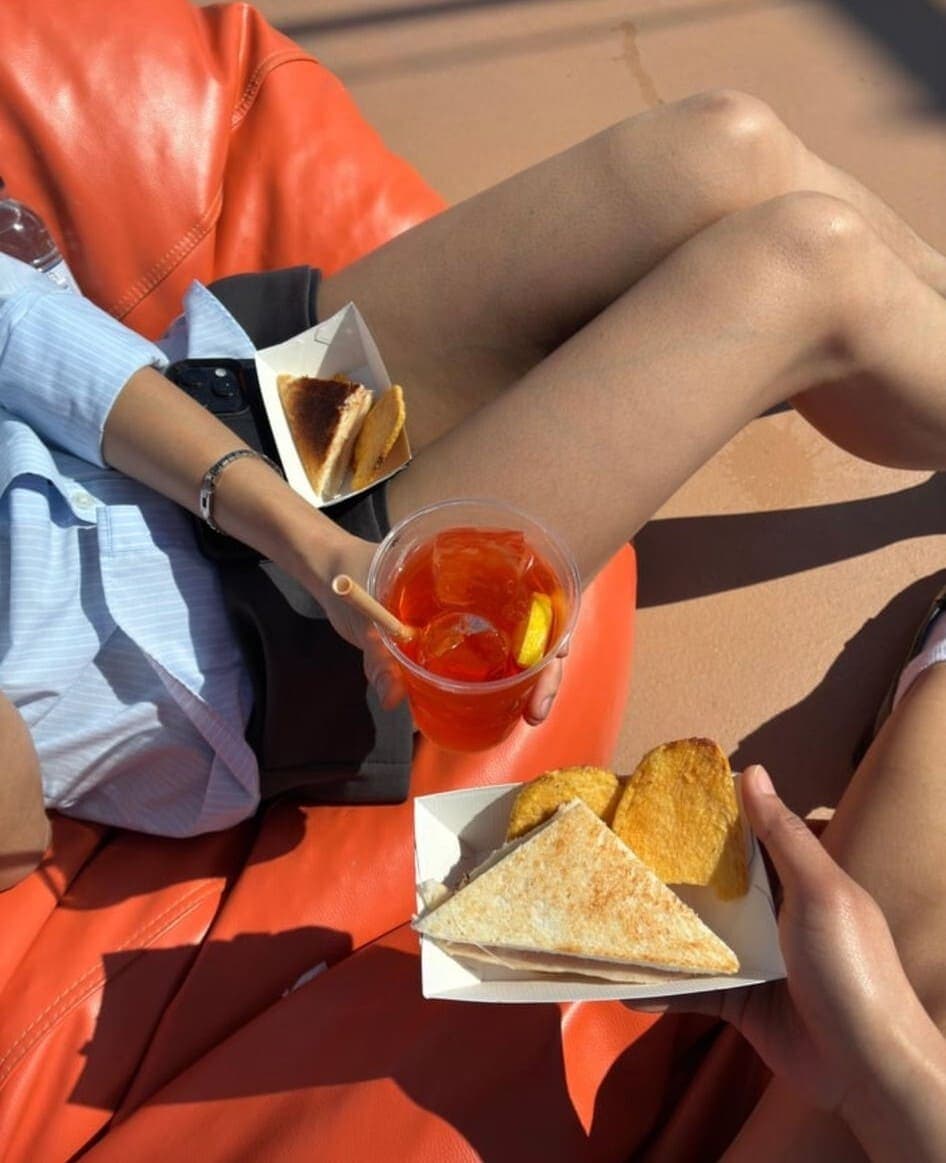 Persona relajada en tumbona naranja con un Aperol Spritz y bocadillo de jamón