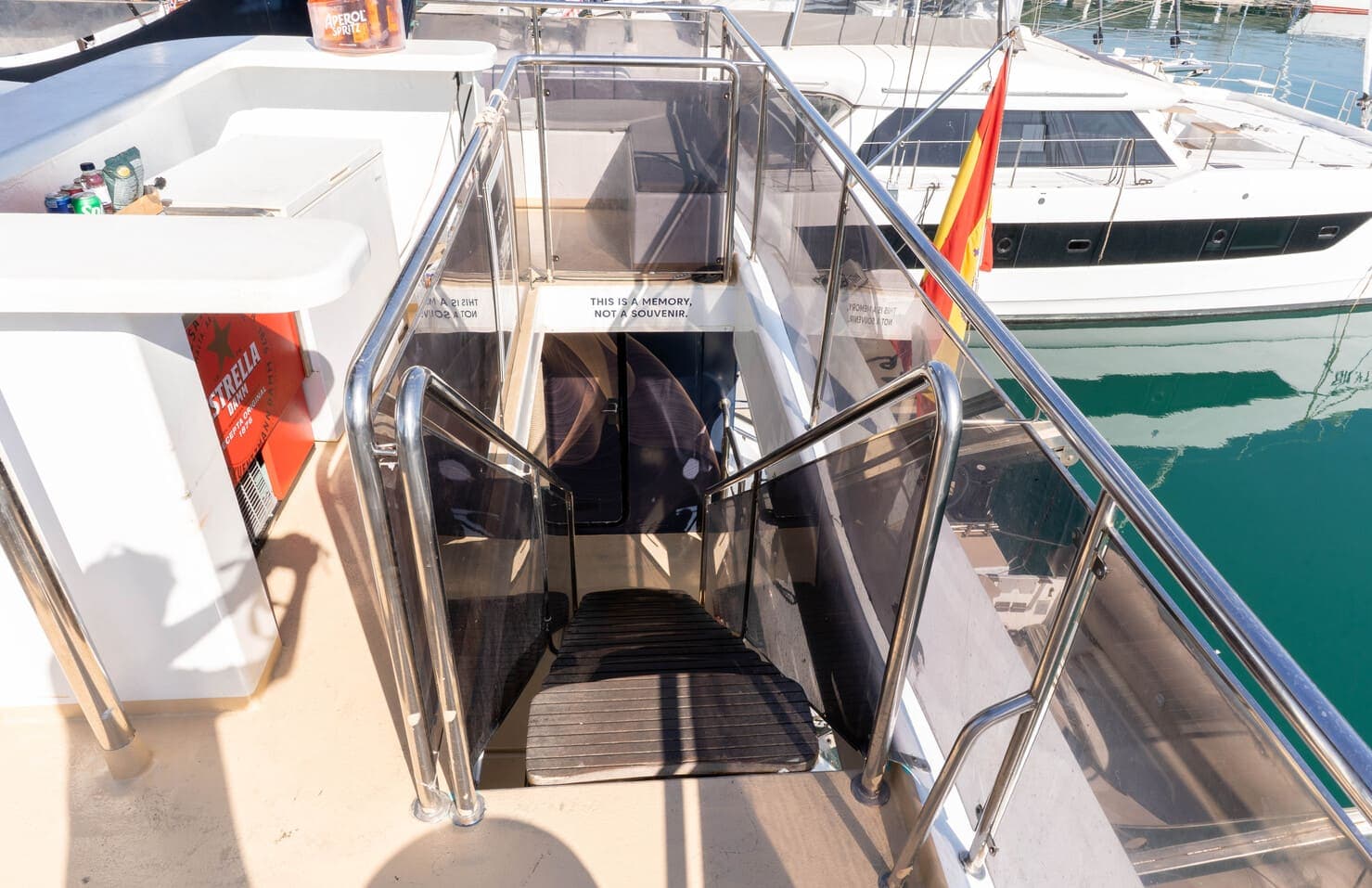 Escalera de acceso con barandillas de cristal en la popa de un catamarán, con un velero al fondo