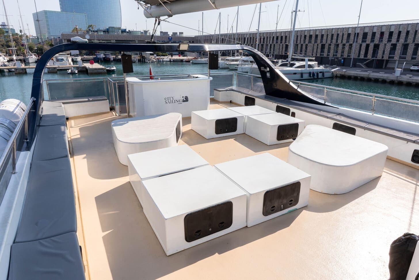Zona de estar en la cubierta de un catamarán con la barra al fondo y el hotel W de Barcelona