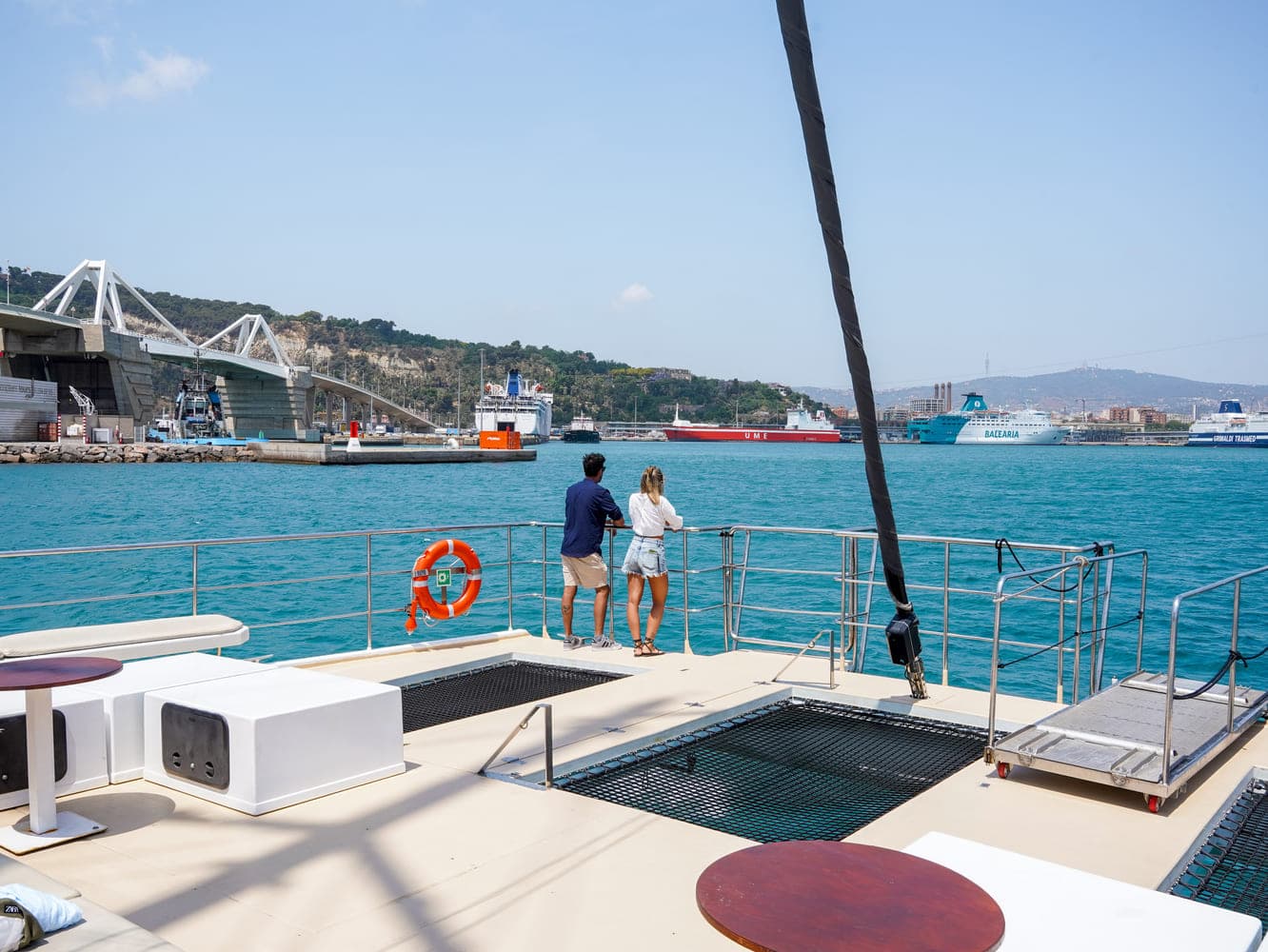 Pareja apoyada en la barandilla de proa de un catamarán observando el puente y un ferry en la bahía