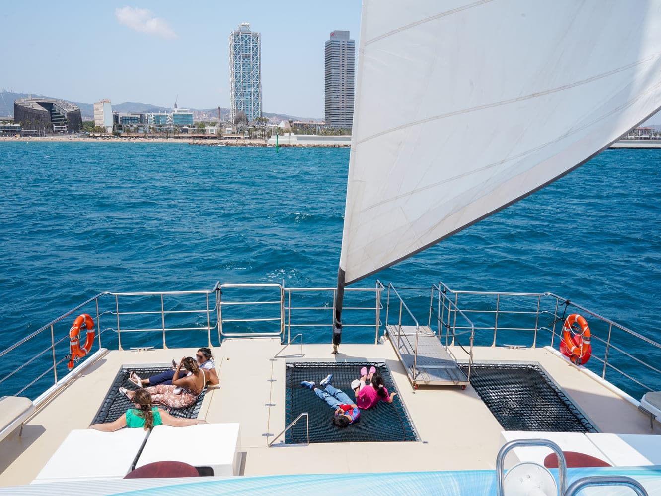 Familias tumbadas en las redes de proa de un catamarán con velas izadas y los rascacielos de Barcelona al fondo