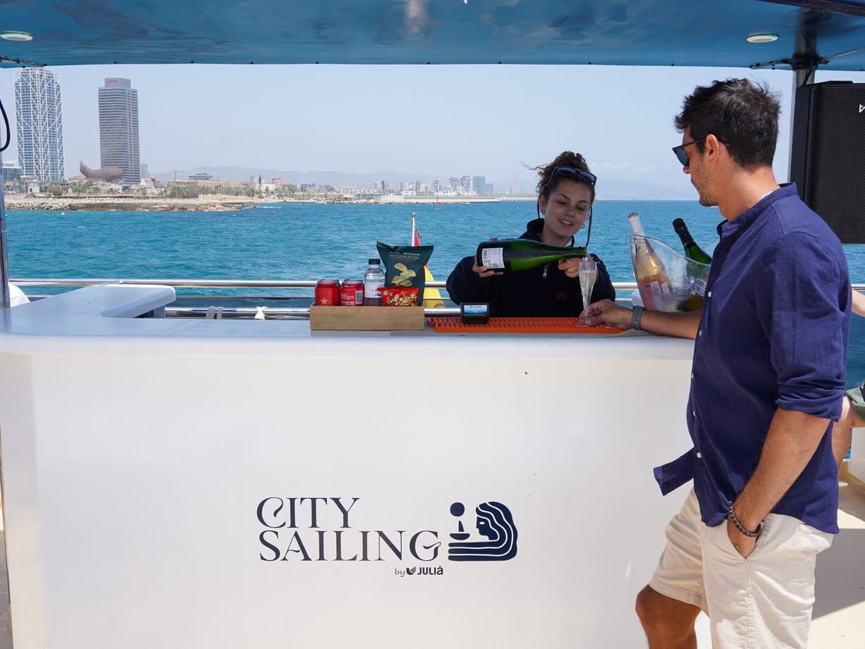 Camarera sirviendo cava en la barra de un catamarán con la costa de Barcelona al fondo